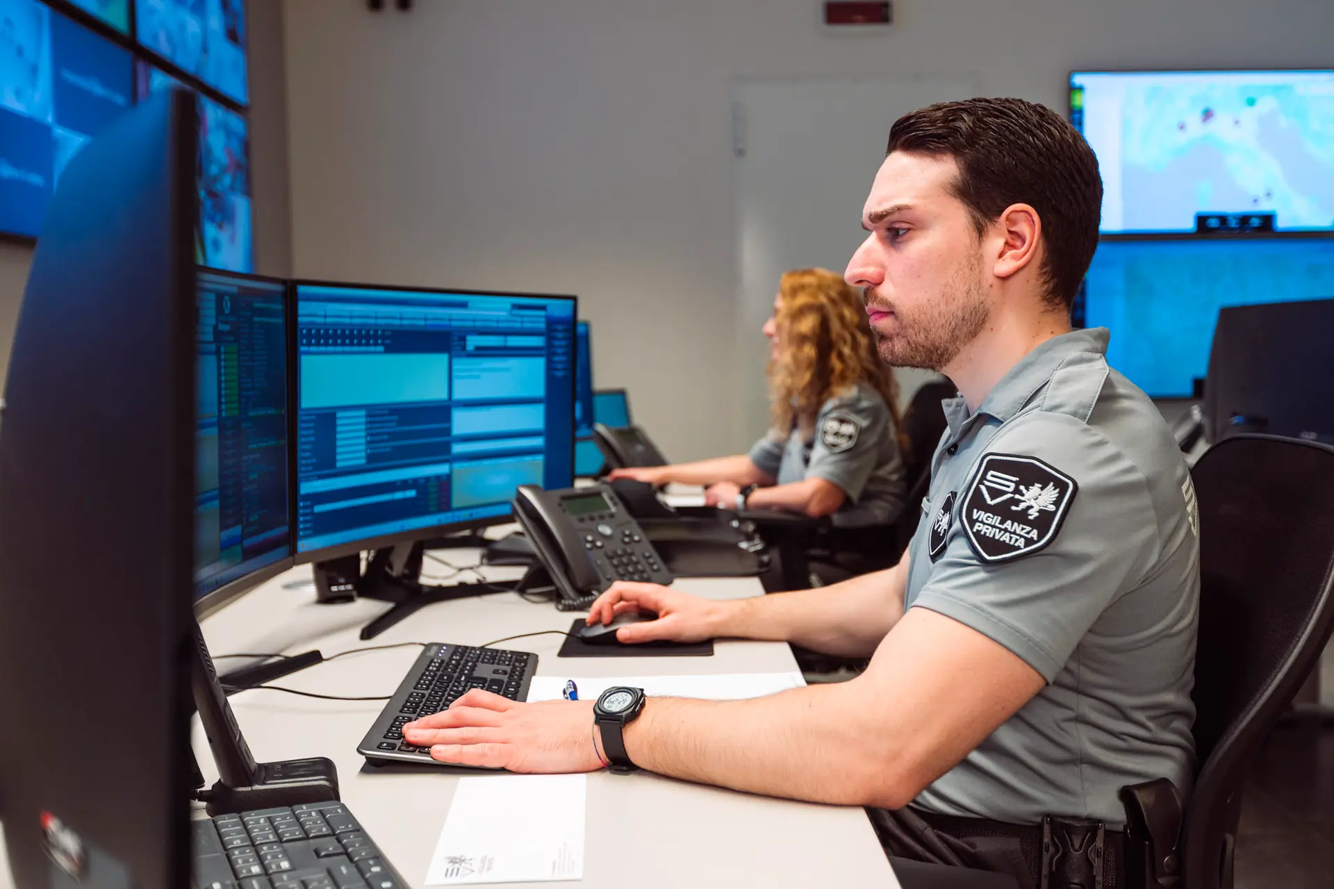 Operatori di vigilanza privata monitorano sistemi di sicurezza da una centrale operativa, davanti a più schermi e telefoni.