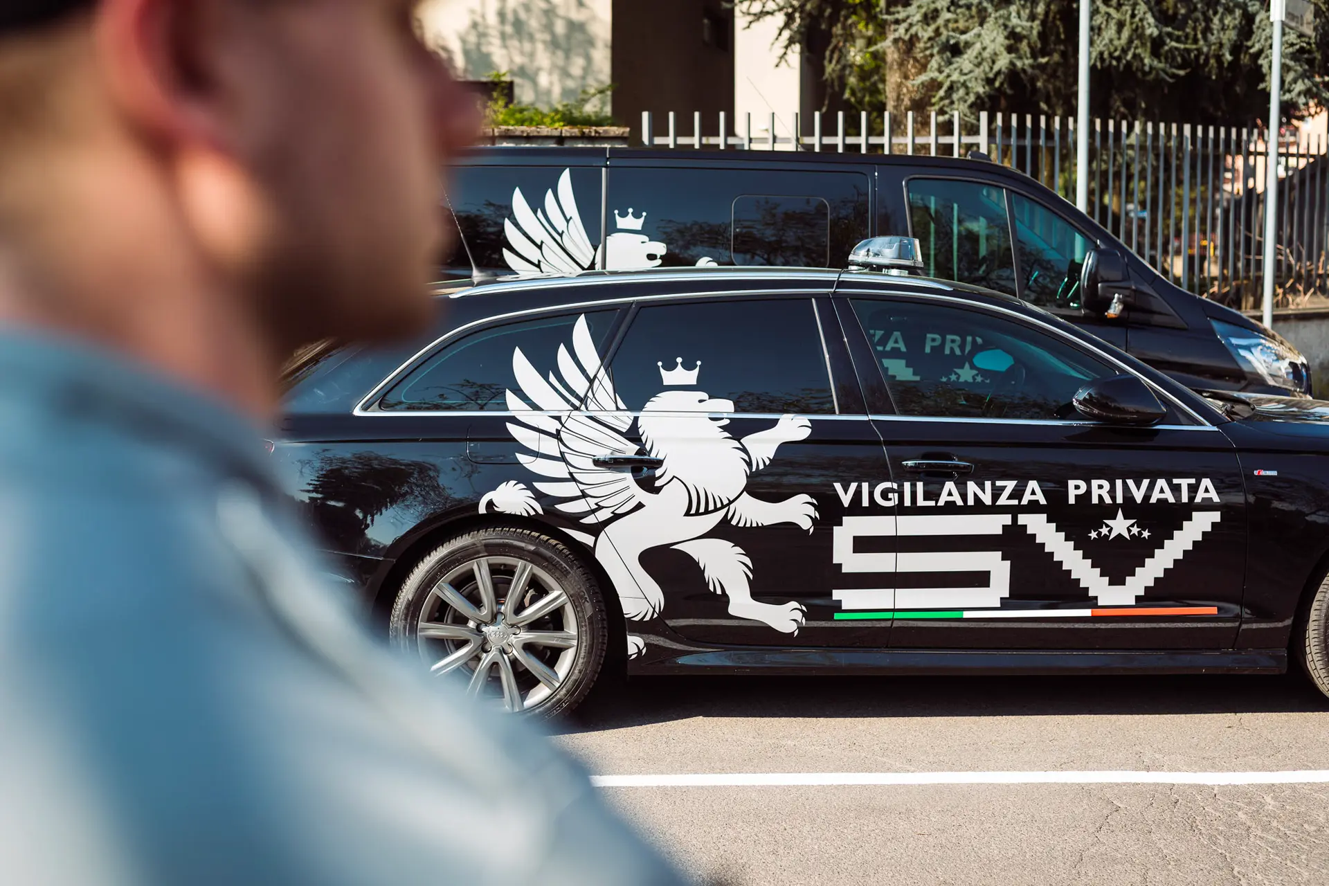 Auto di vigilanza nera con logo SV e leone alato, sullo sfondo di un parcheggio, agente in primo piano sfocato.