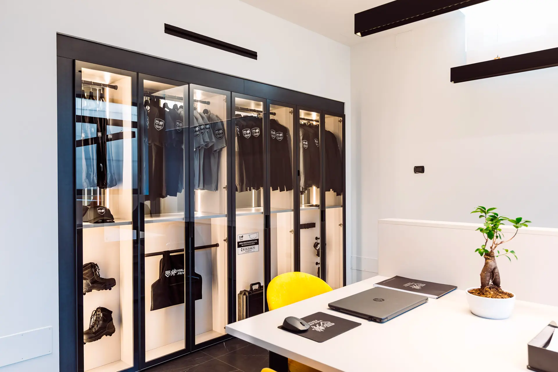 Armadio vetrato con uniformi da vigilanza privata, cappellini, stivali e accessori ordinati per il servizio.
