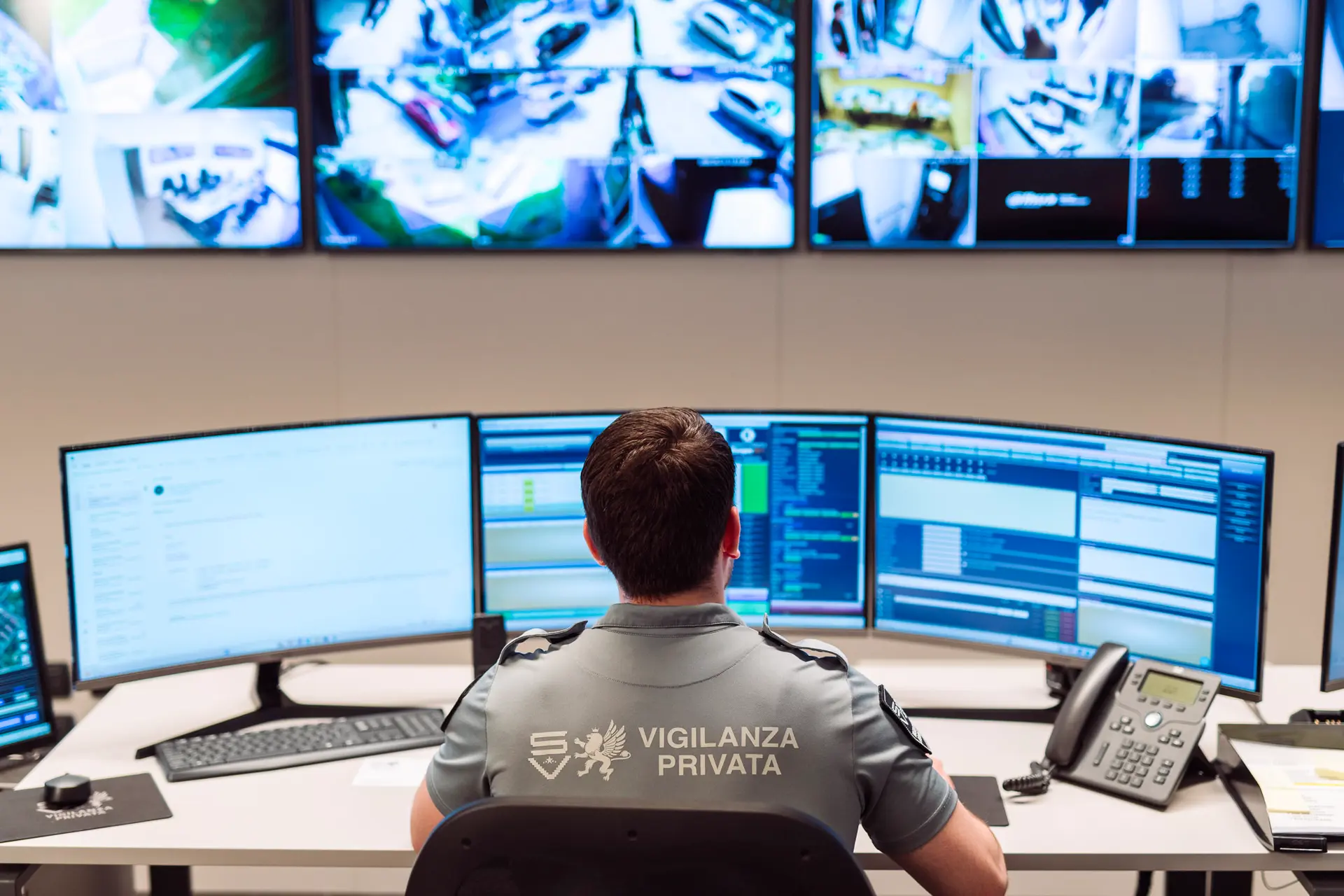 Agente di vigilanza alla scrivania in control room, monitora più schermi con software di sorveglianza attivi.