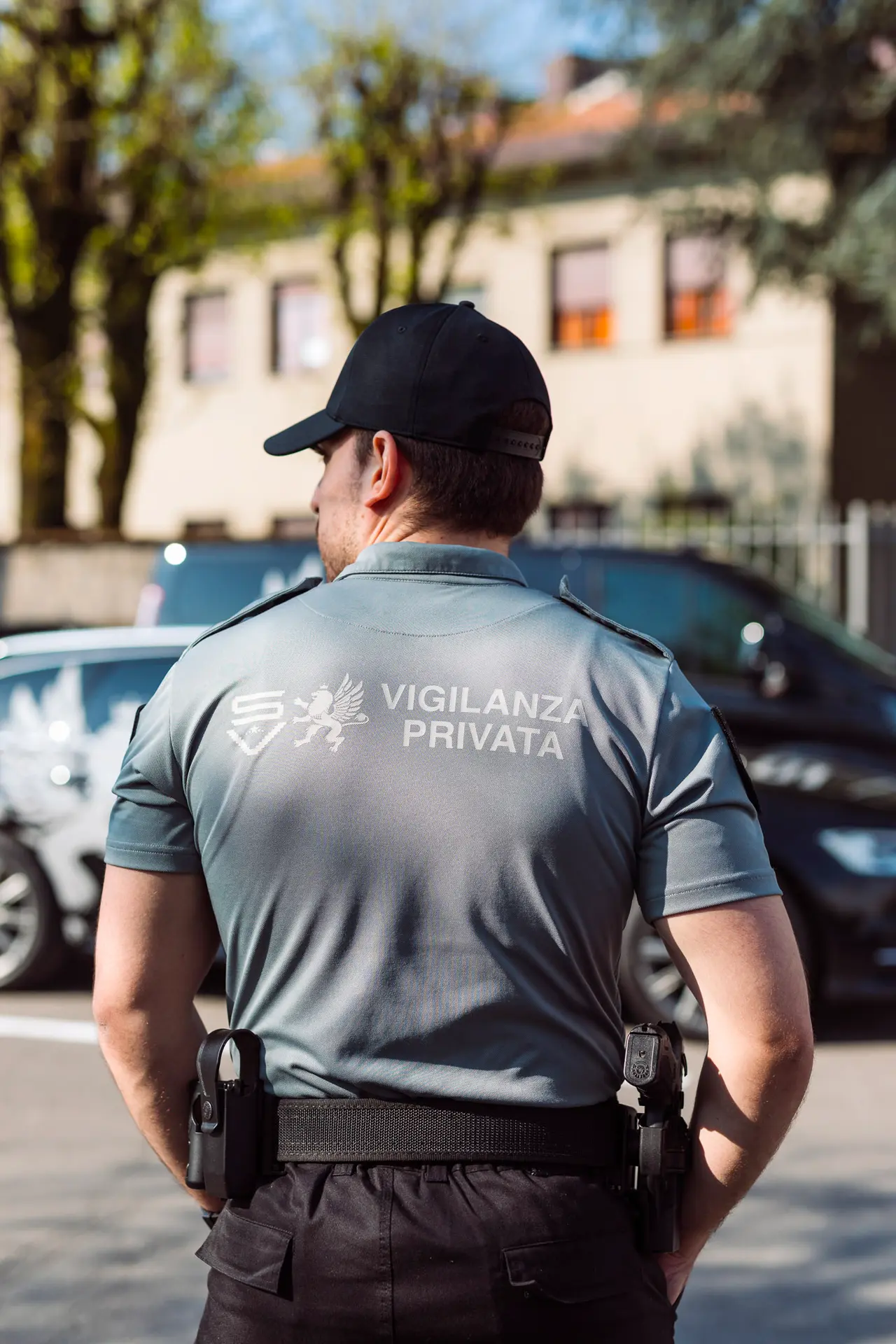 Guardia giurata vista di spalle con divisa grigia SV, in piedi all’aperto vicino a mezzi aziendali parcheggiati.