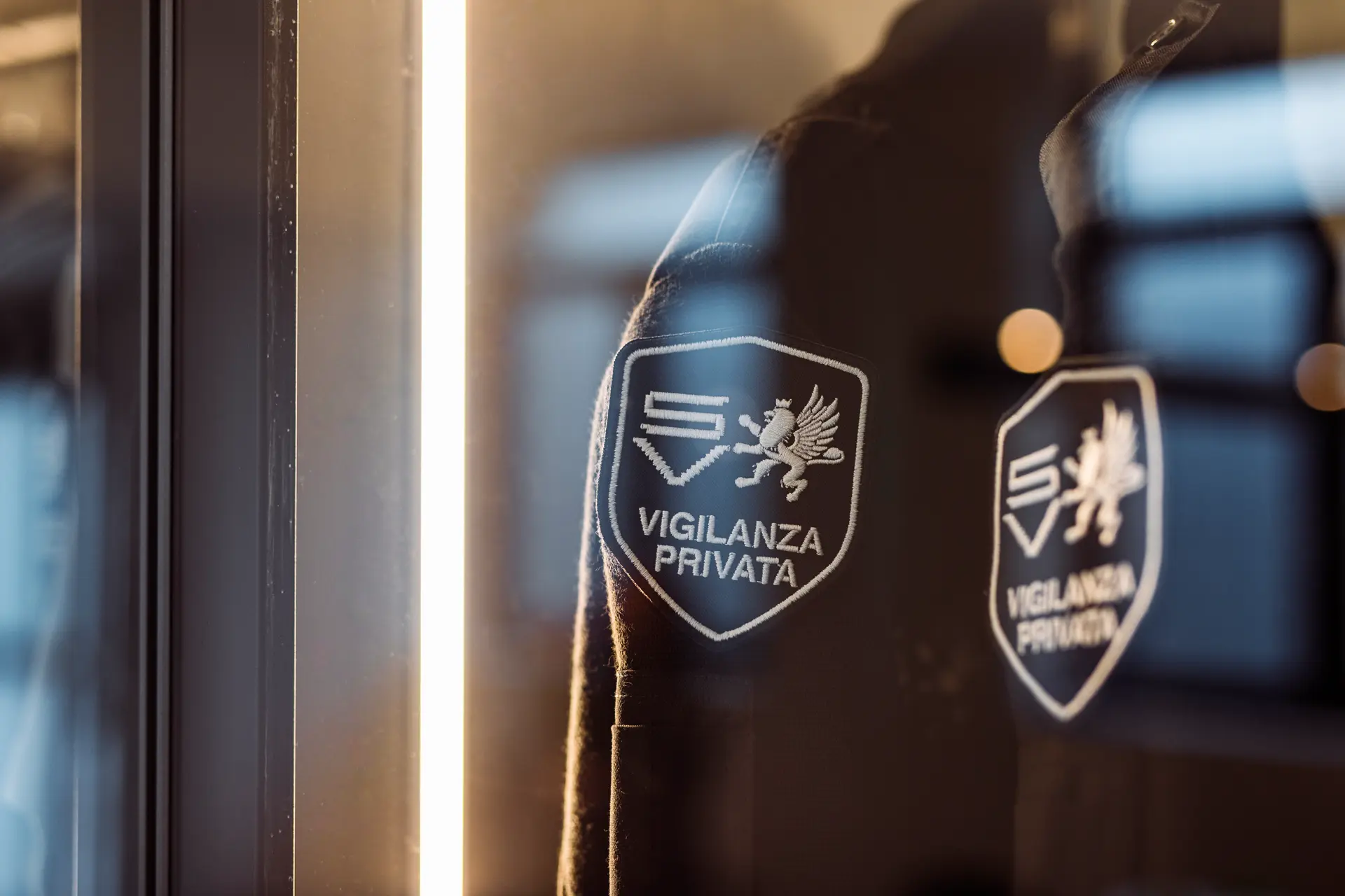 Dettaglio di una divisa da vigilanza privata dietro una vetrina illuminata, con patch ricamata SV e simbolo del leone alato visibile anche nel riflesso sul vetro, simbolo di sicurezza e autorevolezza nel settore della sorveglianza.