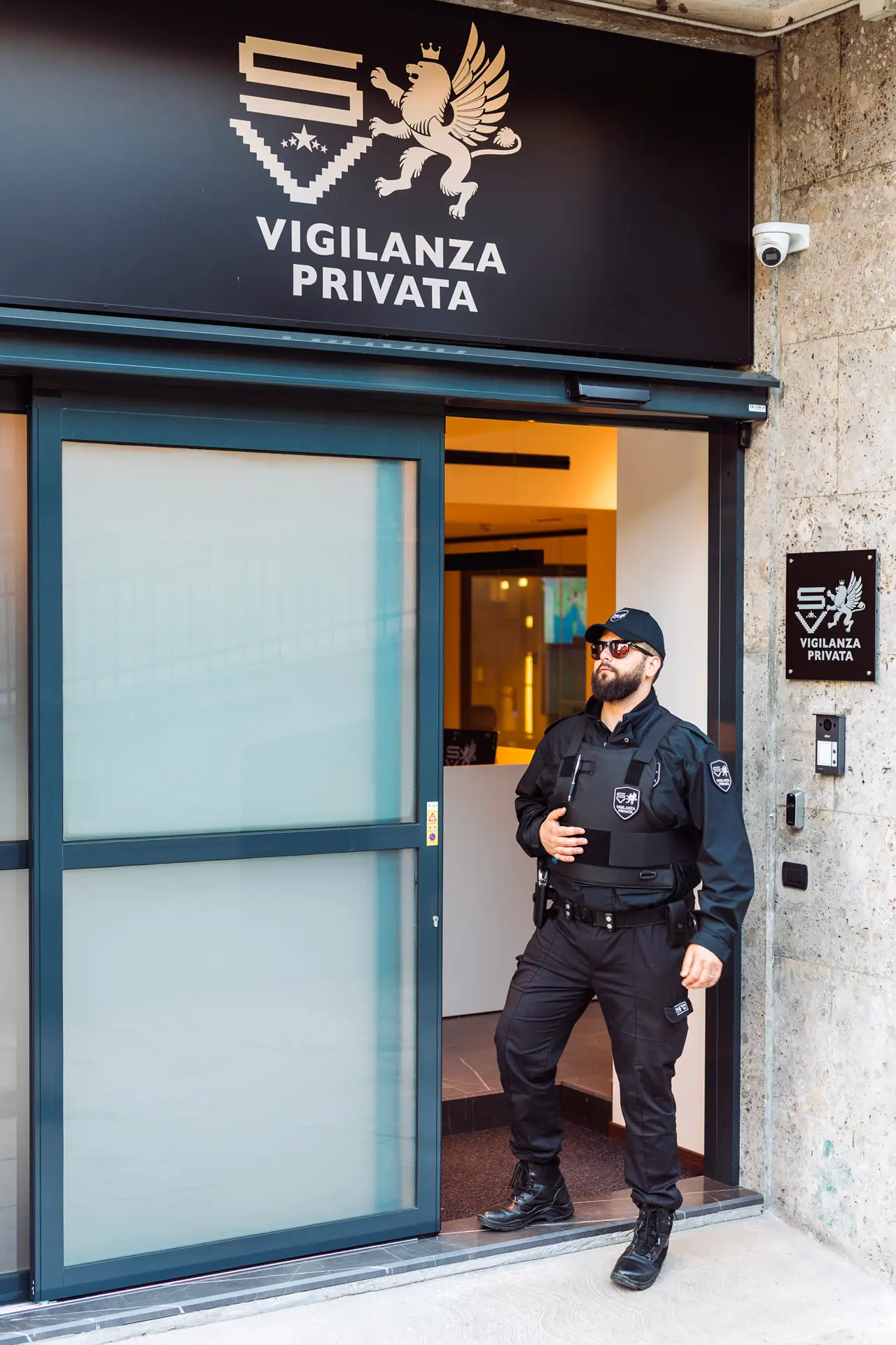 Agente di vigilanza privata esce dalla sede aziendale SV con passo deciso, indossa uniforme nera e giubbotto antiproiettile. Sullo sfondo l’insegna con leone alato e logo aziendale, a sottolineare autorità, sicurezza e prontezza operativa.