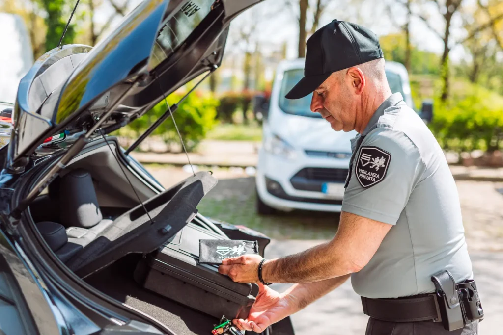 Guardia giurata in uniforme SV apre il bagagliaio dell’auto di servizio e maneggia una valigetta con logo aziendale. Sullo sfondo un furgone e un ambiente alberato, segno di professionalità e preparazione operativa sul campo.