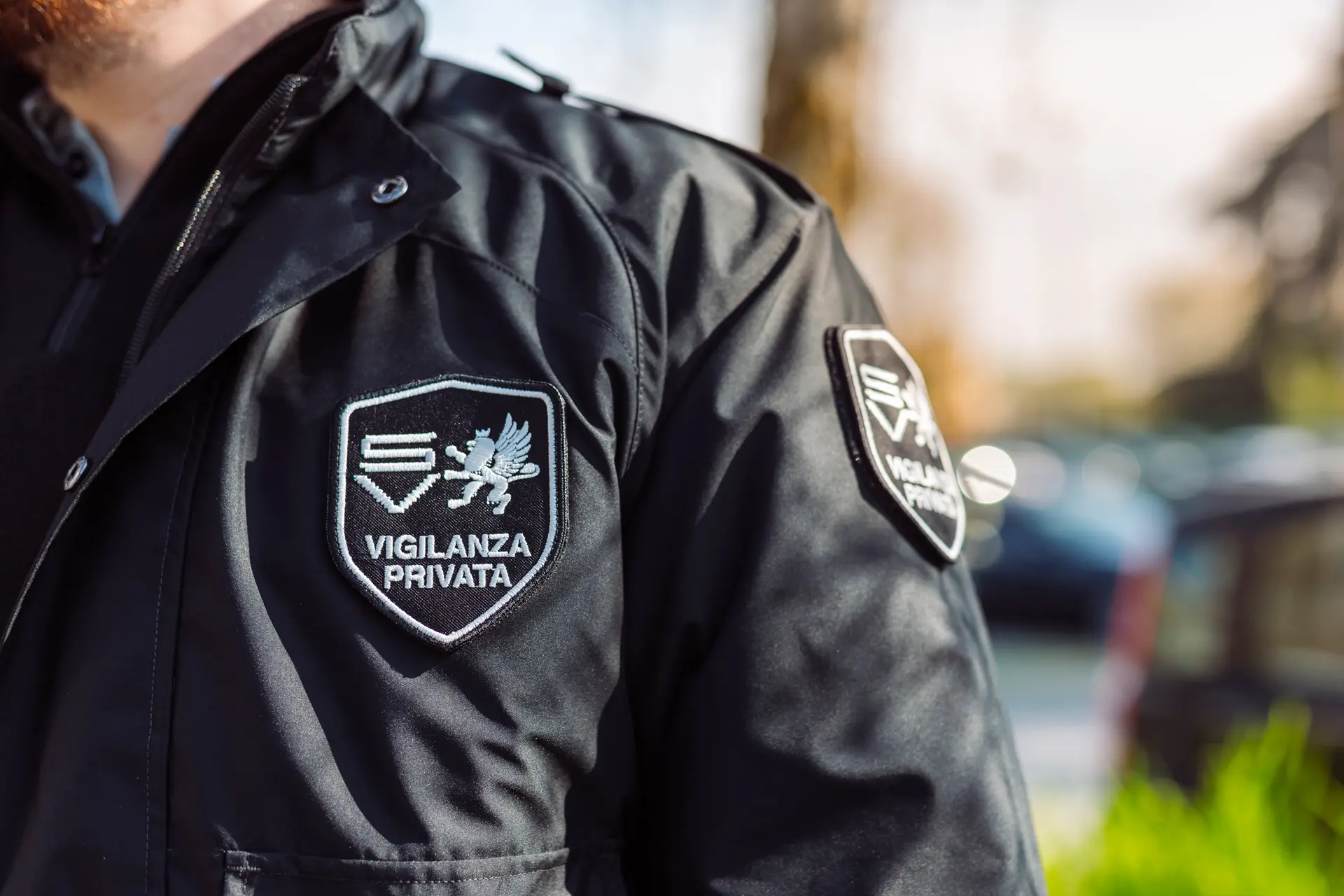 Dettaglio della giacca di un agente di sicurezza privata con logo ricamato, sullo sfondo un parcheggio sfocato.