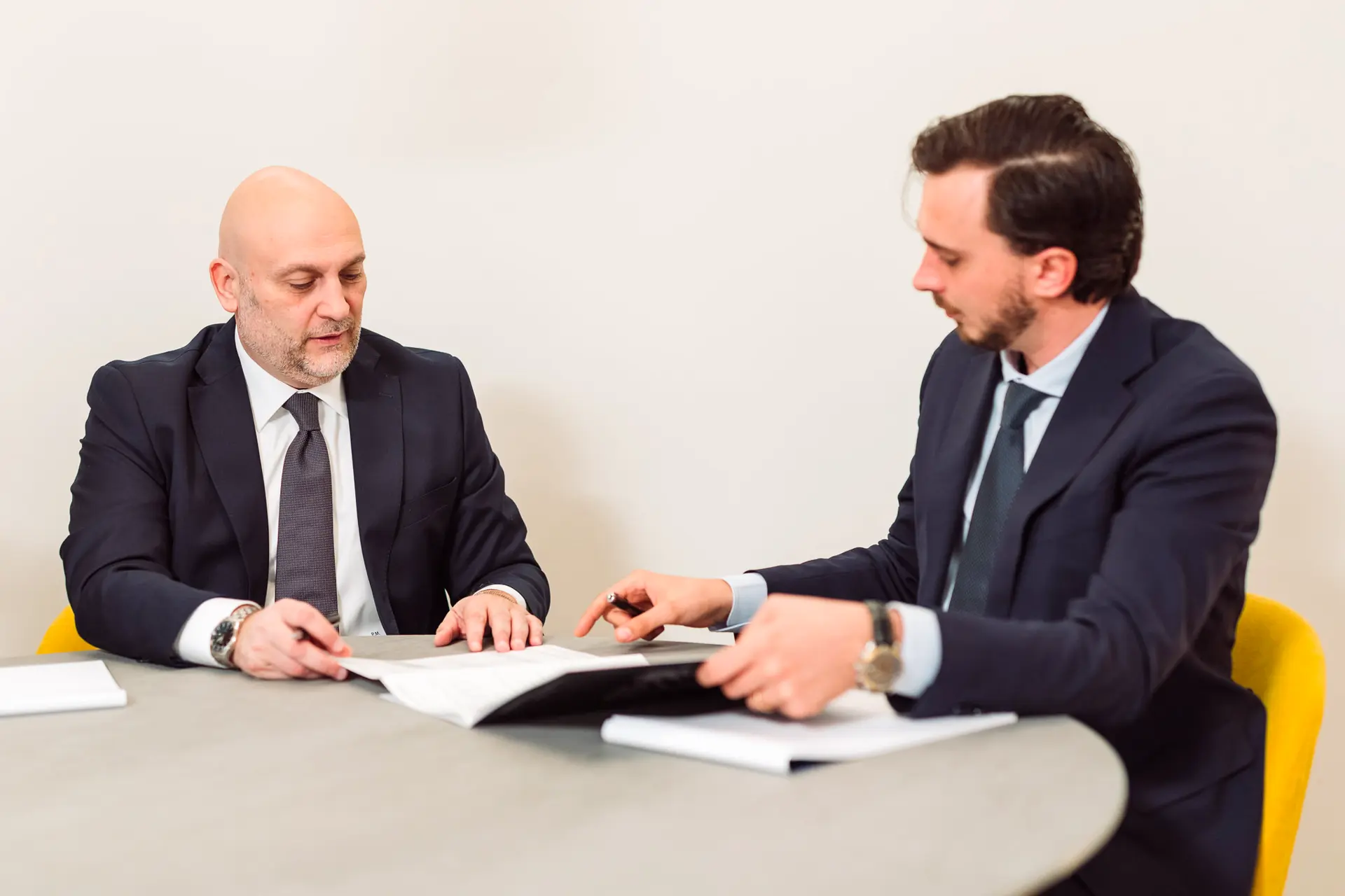 Due uomini in abito elegante discutono seduti a un tavolo tondo, consultando insieme dei documenti stampati.