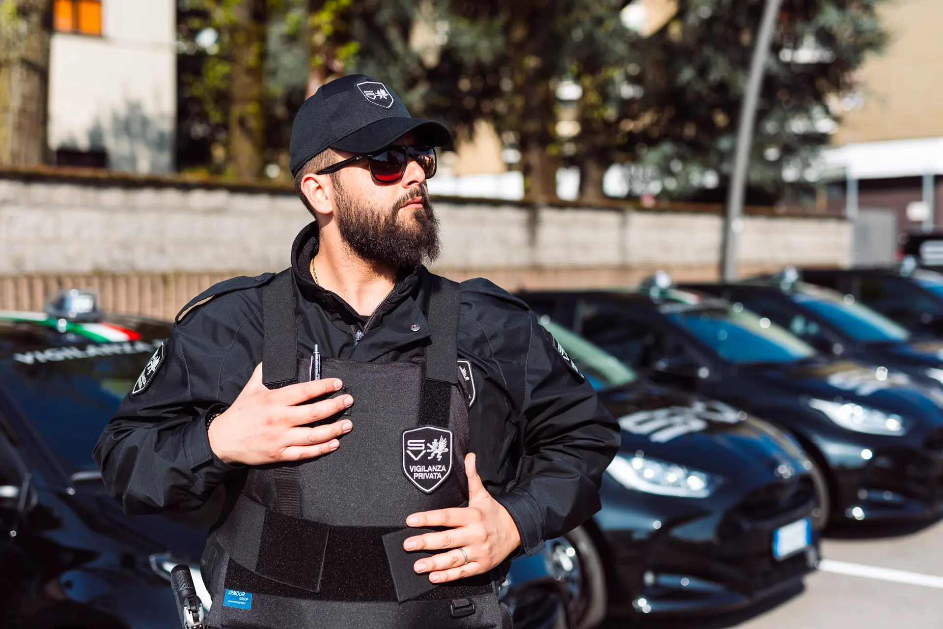 Guardia giurata con barba e occhiali da sole indossa giubbotto antiproiettile in un parcheggio con auto aziendali.