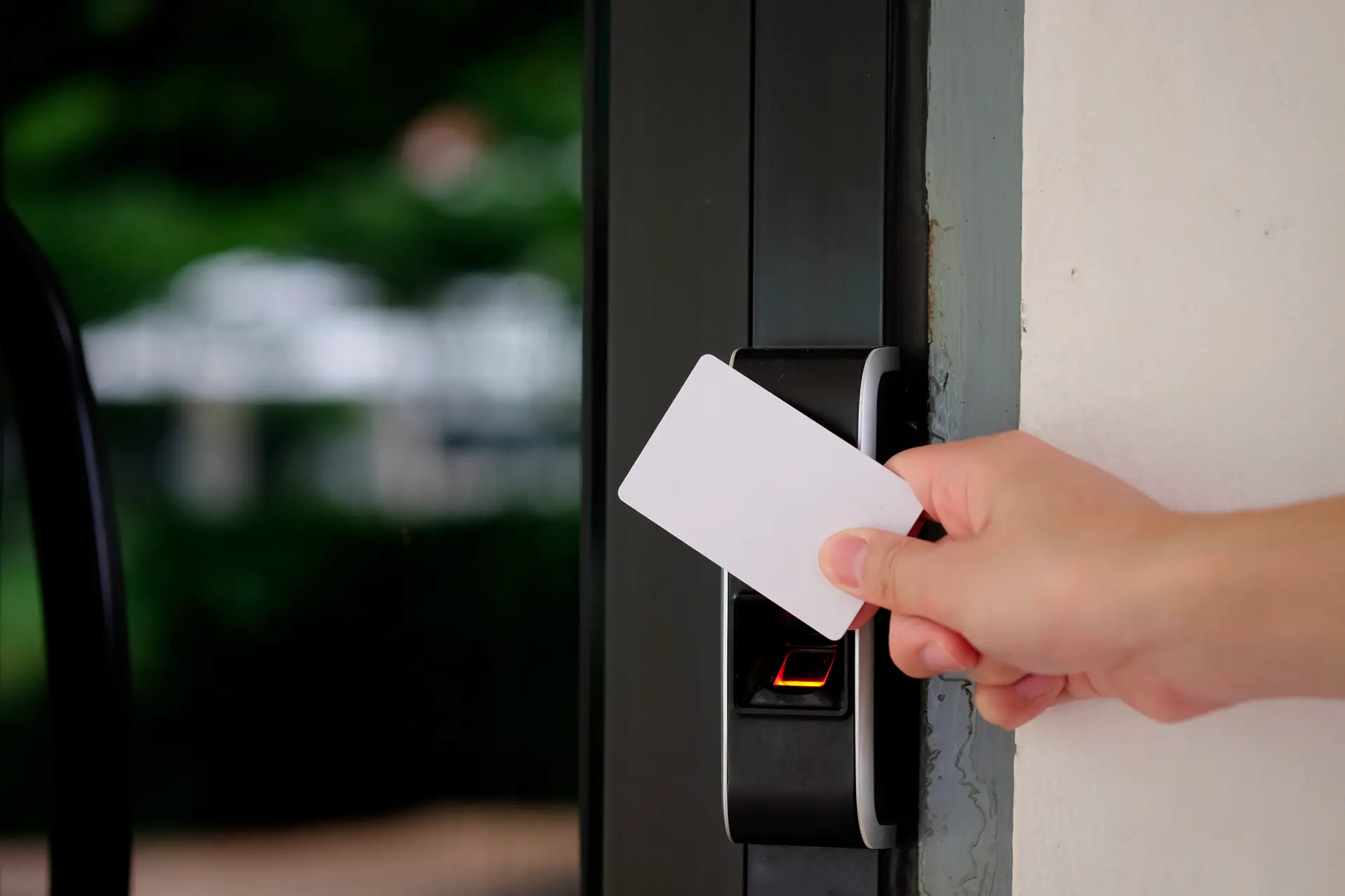 Un dispositivo di accesso elettronico su una porta, con una mano che tiene una carta bianca. Il lettore di accesso si illumina di rosso mentre la carta viene avvicinata. L’ambiente esterno è parzialmente visibile attraverso la porta.