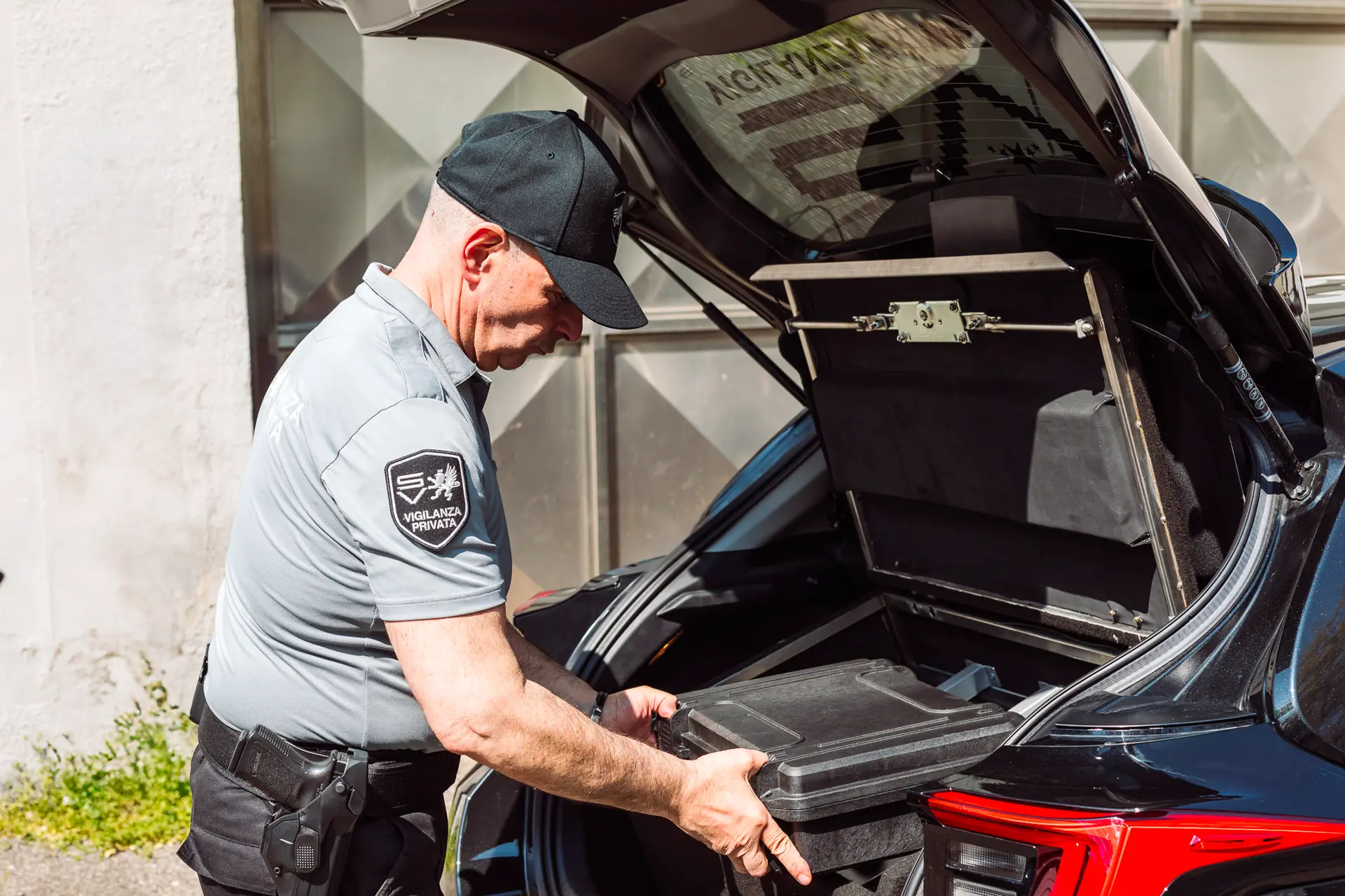 Guardia giurata in uniforme apre il bagagliaio di un’auto e ispeziona una valigetta nera con attenzione.