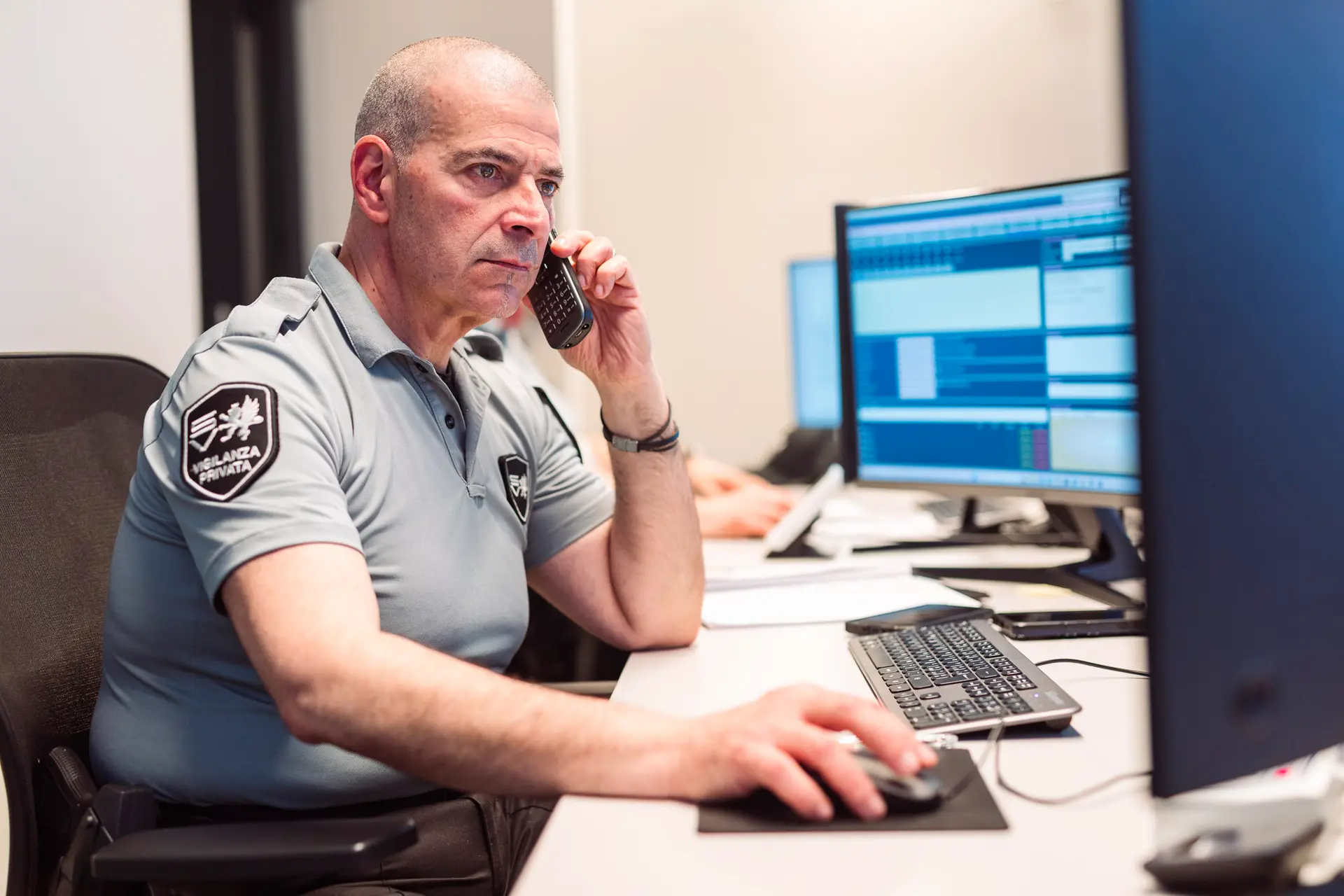 Operatore di vigilanza in uniforme al centralino monitora allarmi su più schermi mentre parla al telefono e usa il mouse.