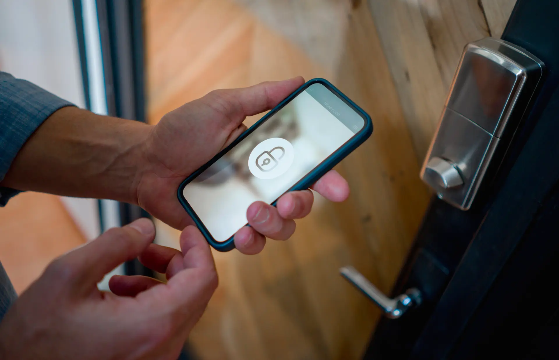 Una mano tiene un telefono cellulare con l'icona di un lucchetto sullo schermo, mentre si prepara a sbloccare una porta. Il lettore della porta è visibile accanto al telefono, e l'ambiente circostante è parzialmente sfocato.
