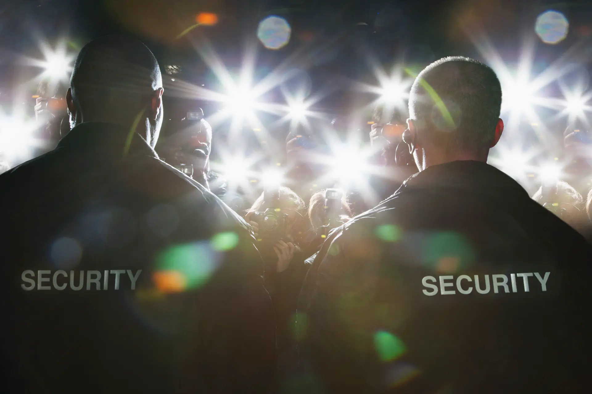 Due agenti di sicurezza indossano giacche con la scritta 'SECURITY' mentre proteggono una persona famosa. Le luci dei flash delle fotocamere lampeggiano intensamente mentre la folla si avvicina.