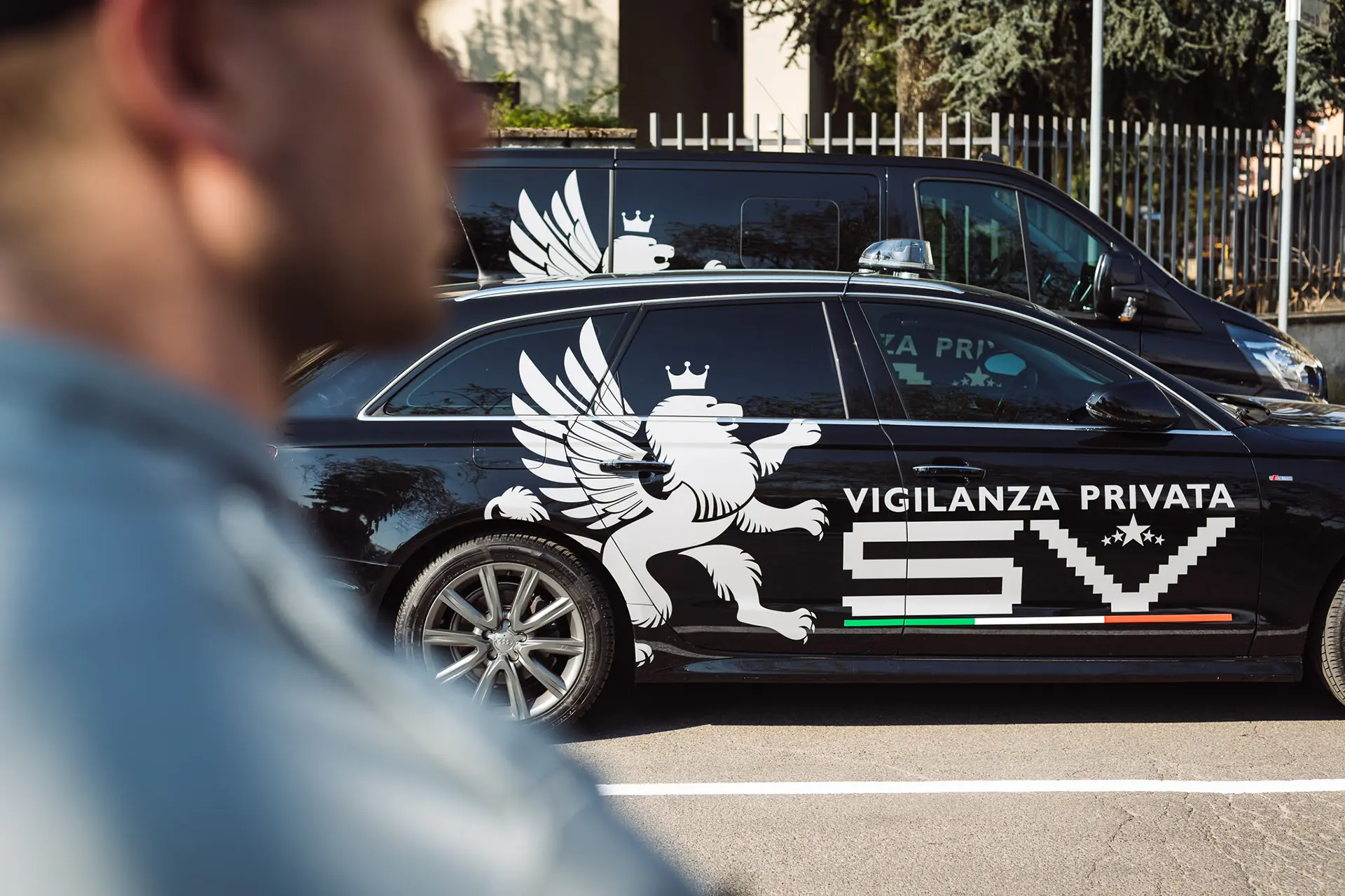 Auto nera con logo SV Vigilanza Privata e grifone bianco stilizzato sul fianco, in primo piano un agente sfocato di spalle.