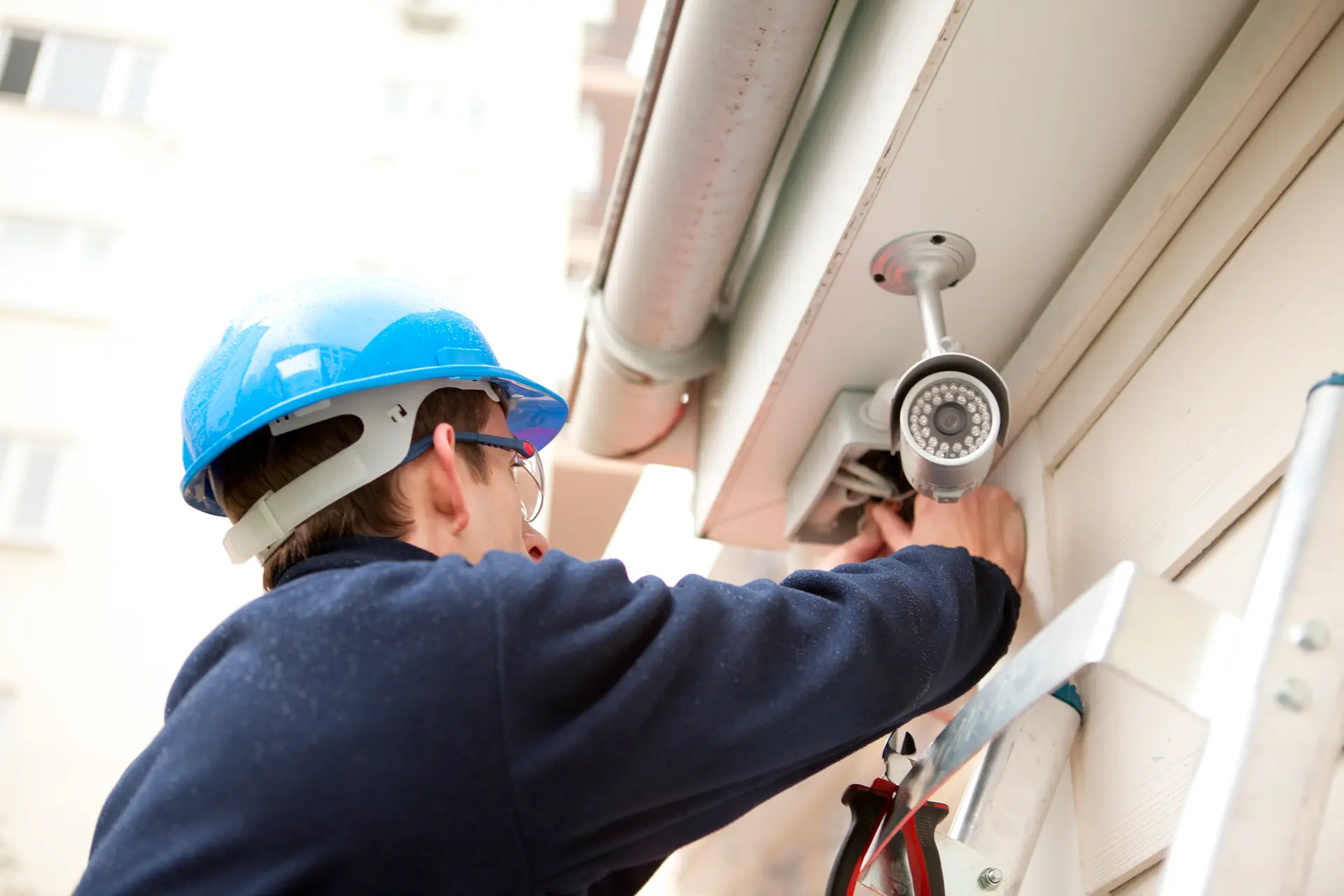 Un uomo con casco blu e occhiali sta sistemando una telecamera di sorveglianza su una parete esterna di un edificio. È in piedi su una scala e appare concentrato sul compito. La telecamera è montata sull'angolo dell'edificio, con un ambiente luminoso e finestre visibili sullo sfondo.
