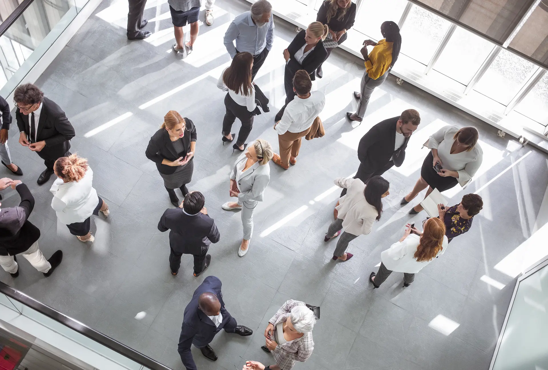 Un gruppo di professionisti, uomini e donne, sono in piedi in un ufficio o sala conferenze. Indossano abiti formali e sono impegnati in conversazioni. La luce è naturale, con la luce solare che entra attraverso ampie finestre, proiettando ombre sul pavimento.