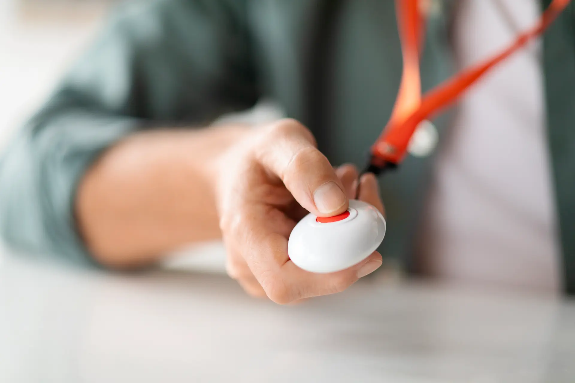 Un primo piano di un pulsante di emergenza bianco tenuto in mano da una persona, con un laccetto arancione attaccato. L'attenzione è sul centro rosso del pulsante, segnalando la sua importanza per una risposta d'emergenza. Lo sfondo è sfocato, mettendo in evidenza la mano e il pulsante.