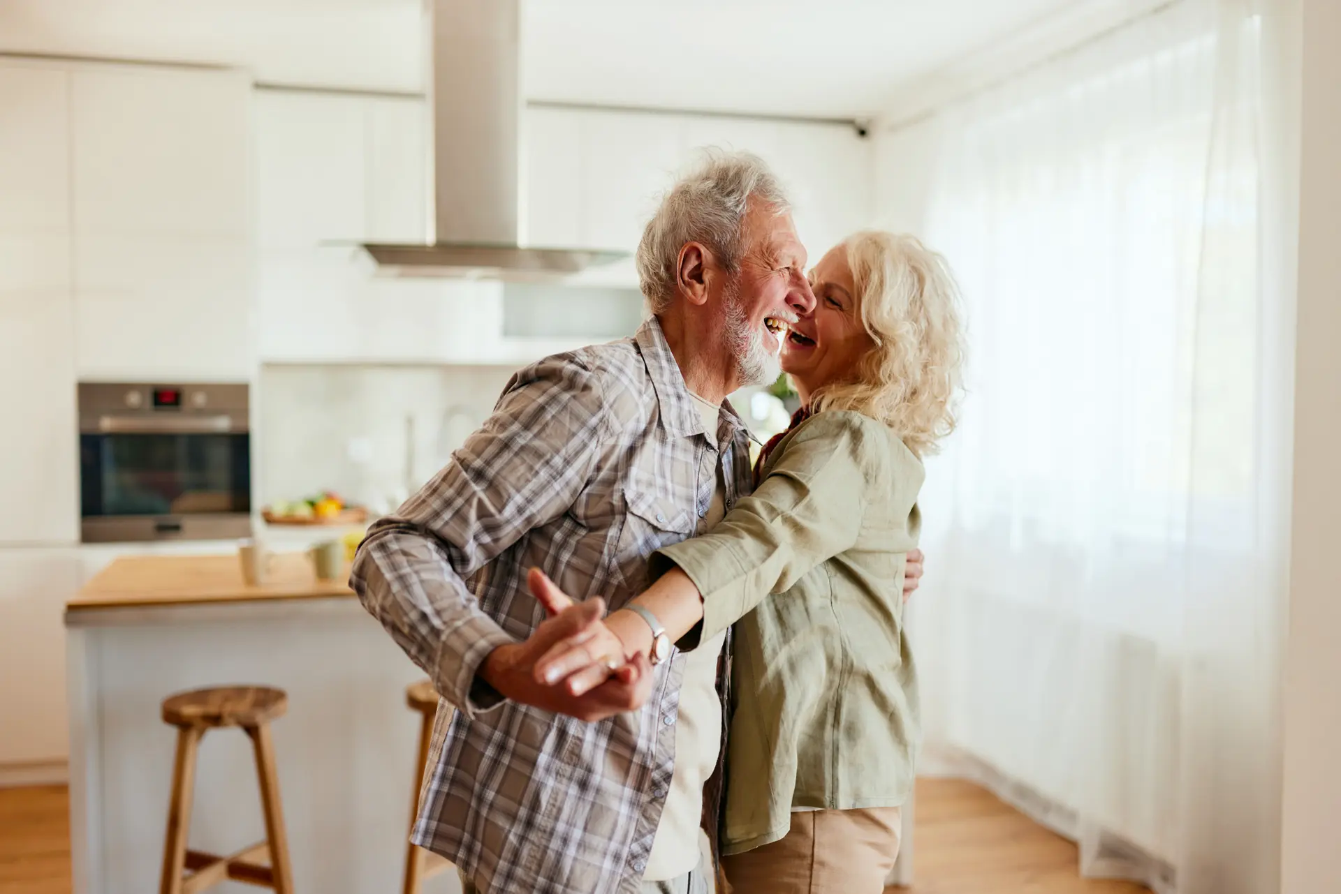 Una coppia di anziani, entrambi sorridenti, stanno ballando insieme nella loro cucina. La donna ha i capelli corti e ricci, mentre l'uomo ha la barba grigia. La cucina è luminosa, con la luce che entra da grandi finestre, creando un'atmosfera calda e gioiosa.