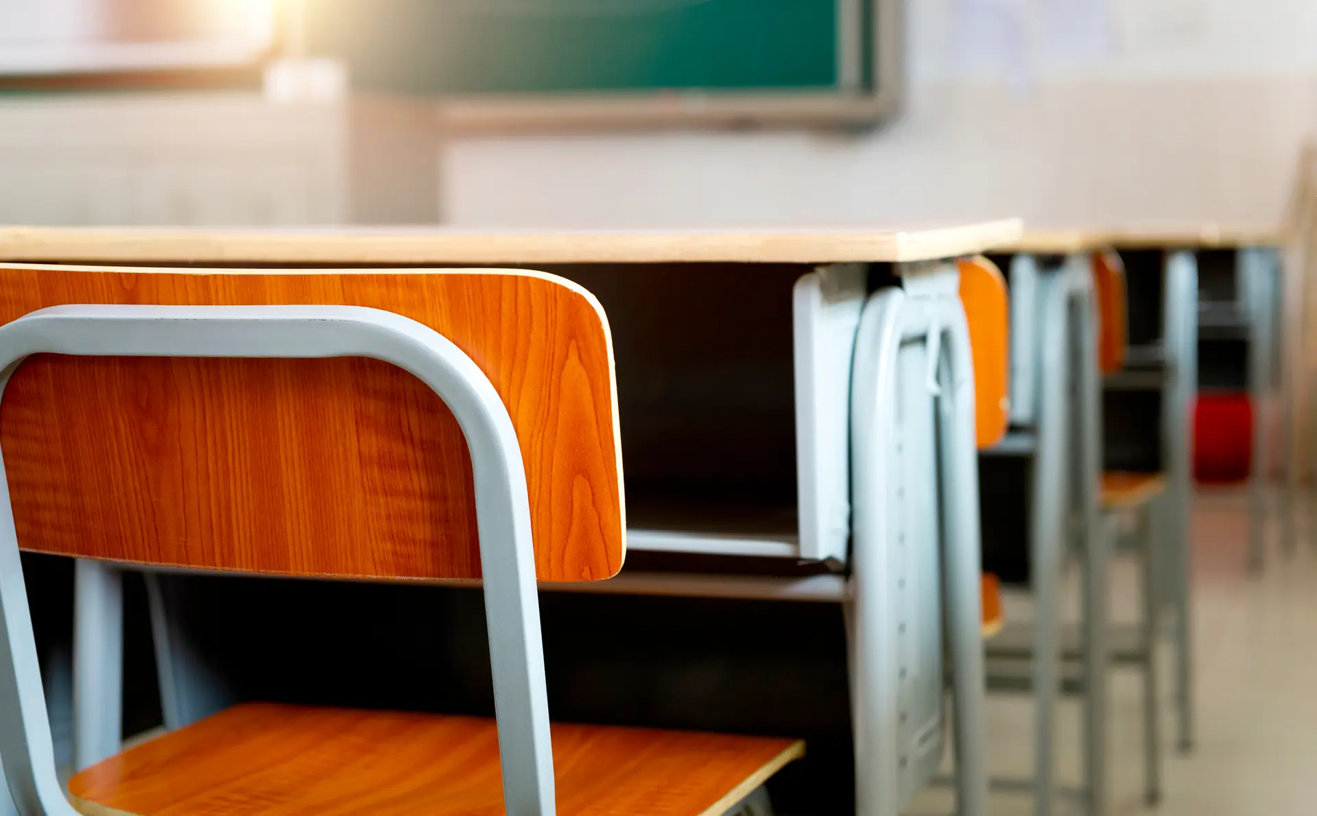 Un primo piano di sedie e banchi in una classe. Le sedie sono in metallo e legno, con la luce solare che entra attraverso una finestra sullo sfondo. L'ambiente della classe è invitante ma organizzato, con un senso di ordine.