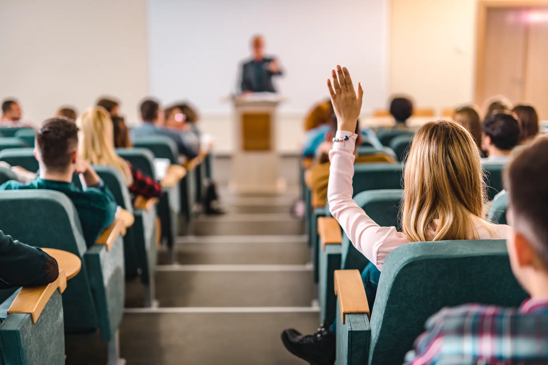 Una sala conferenze con file di sedie occupate da studenti. Una donna nella fila anteriore alza la mano per fare una domanda. Il professore, sfocato sullo sfondo, sta parlando alla classe. L'ambiente suggerisce una collaborazione attiva in un ambiente di apprendimento moderno.